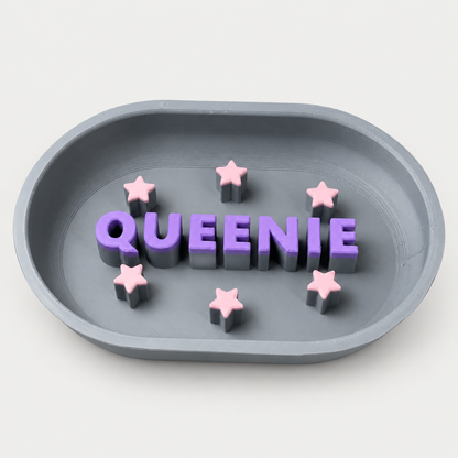 Grauer personalisierter Slowfeeder QUEENIE mit violetten Buchstaben und rosa Sternen auf neutralgrauem Hintergrund